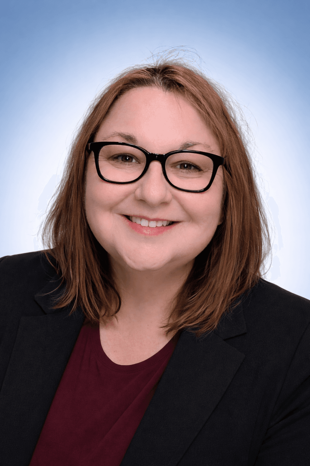 Femme souriante aux cheveux roux et lunettes noires, portant un blazer sur fond bleu clair.